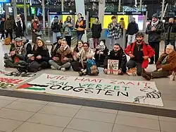 Kleine sit-in op Utrecht Centraal Station. Op de achtergrond zitten en staan pro-Palestijnse demonstranten, op de voorgrond ligt een spandoek met de tekst "Wie haat zaait zal verzet oogsten" en een kleine getekende Palestijnse vlag. Daarachter een kleine poster met de tekst "Resistance is justified when people are occupied".