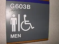 Verenigde Staten — Een bord met verhoogde pictogrammen en brailletekst dat de weg wijst naar een toegankelijk toilet. Het bord is bevestigd op grijphoogte naast een deur in een openbaar gebouw, conform ADA-normen.