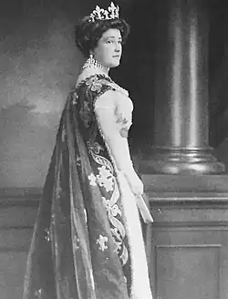 Donkerharige vrouw in begin 20ste-eeuwse kleding, foto en profil, met waaier in haar rechter hand en koninklijke cape versierd met borduursel over haar schouder. Ze draagt grote oorbellen en choker ketting en een diamanten tiara in fleur-de-lys motief.