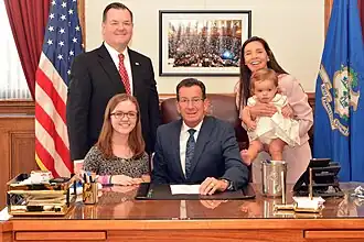 Links onderaan staat een volwassen achondroplaste, rechts houdt een moeder haar baby met achondroplasie vast. Ze poseren met Dannel P. Malloy, gouverneur van de Amerikaanse staat Connecticut, bij de ceremoniële ondertekening van de Wet 15-87 waarin de 'Dwarfism Awareness Month' officieel wordt erkend.