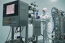 Een persoon in een volledig wit cleanroom-pak, blauwe handschoenen en een gezichtsmasker bedient complexe roestvrijstalen bioprocessingapparatuur in een steriel laboratorium.