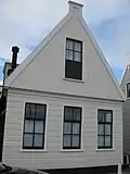 Houten huis met skelet. Puntvormig voorschot, goede pui en roedenverdeling. Gepotdekselde achtergevel met goede roedenverdeling in de vensters en een deurtje uit derde kwart 18e eeuw
