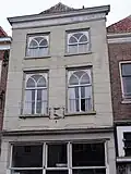 Huis met rood schilddak en gebosseerd grijsgepleisterde lijstgevel, ramen met waaierzwikken; gevelsteen in de vorm van een cartouche waarin een pauw. Winkel