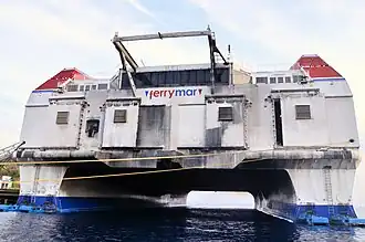 De achterkant beeldvullend, van onderaf gezien, met een blik tussen de rompen van de catamaran door. Het witte achterschip zit vol zwarte vegen en plekken van roet uit de dieselmotoren. Beide foto's zijn eind 2012 genomen bij Curaçao, waar het schip in 2011 opgelegd was.