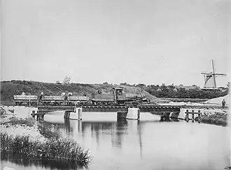 Een werktrein rijdt over een kraanbrug over de gracht van de linie bij de oostelijke redoute, gezien vanaf het Sterrebos. Op de achtergrond een van de lunetten of forten (1868)