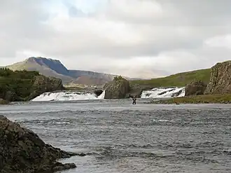 De Laxfoss. Achter het linkerdeel van de waterval zijn de bebouwing van Bifröst en de Grábrók herkenbaar, achter het rechterdeel ligt Baula deels in de wolken verscholen.