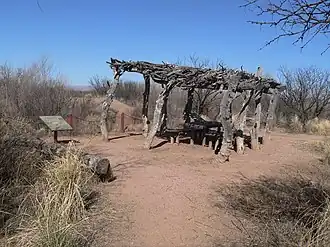 Een open structuur met een dak van takken opgehouden door verticale boomstammen (Ramada, interpretive trail), Murray Springs Clovis-site