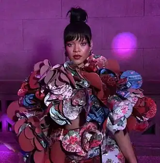 Een ingezoomde foto van Rihanna op het Met Gala van 2017. Ze is gekleed in een kleurrijke, driedimensionale jurk ontworpen door Rei Kawakubo voor het modemerk Comme des Garçons. De jurk bestaat uit een reeks overlappende, bloemvormige panelen van stof in felle kleuren en drukke patronen, waaronder rood, roze, blauw en paars. De  stofpanelen creëren een driedimensionaal effect. Rihanna draagt haar haar in een knot en haar oogschaduw, lipstick en blush zijn verschillende paarstinten. De foto is gekropt en toont alleen haar bovenlichaam. De achtergrond is paars.