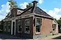 Laag dorpshuis met omlijste ingang, zesruitsvensters en versierde kroonlijst onder schilddak met twee hoekschoorstenen waarop borden