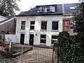 Gepleisterd huis met verdieping en schilddak, haaks op het plein