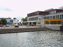 Een gebouw met een pleintje met een kunstwerk ervoor, aan het water